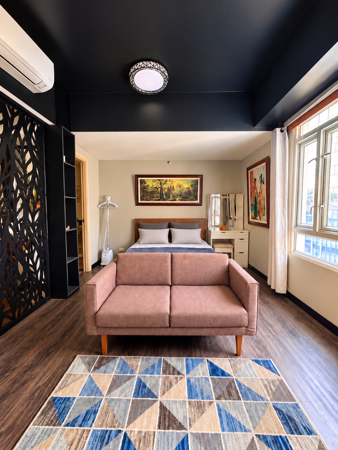 Studio living and sleeping area — dusty-pink sofa, queen bed, vanity, and dark accent ceiling