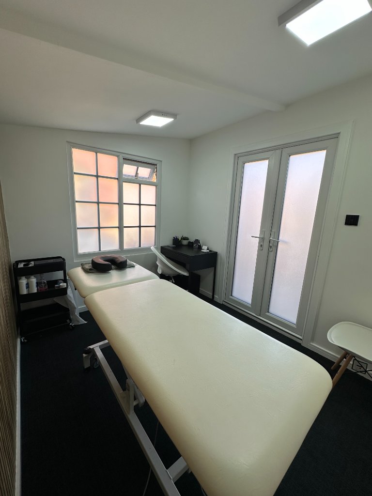 Corner view of the clinic room showing the treatment table, rolling trolley, desk, and frosted French doors