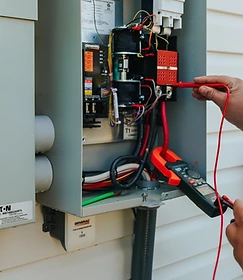 GenPro electrician testing an open electrical panel with a multimeter probe while a Generac surge protector is visible inside the housing