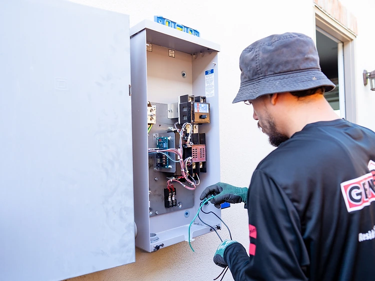 GenPro electrician in a black branded shirt inspecting an open residential electrical panel with a green wire in hand