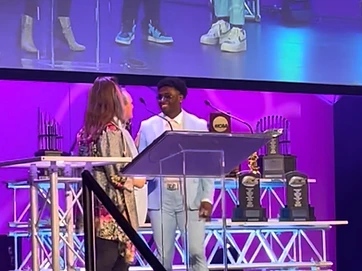 Elijah Green speaking at a podium under purple stage lighting during a live event