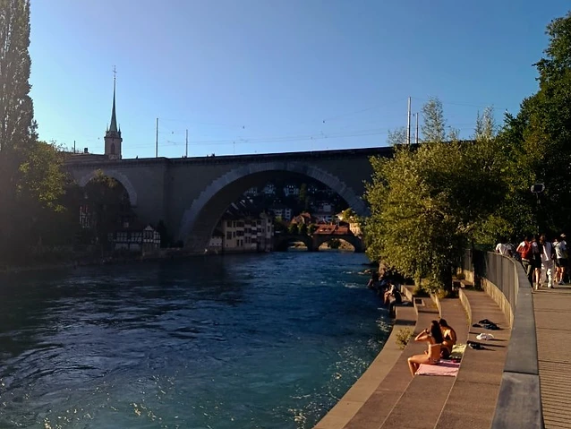 People resting on riverside concrete steps with a large bridge in the distance.