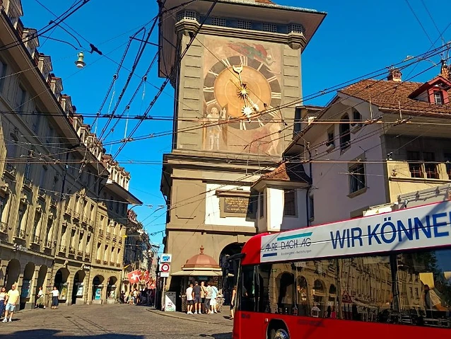 A red tram passes a medieval clock tower on a bustling city street.
