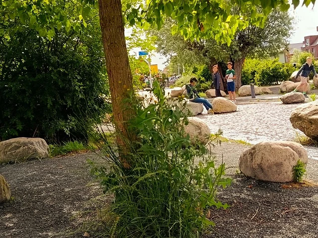 People sit on scattered boulders within a green urban space with gravel paths.