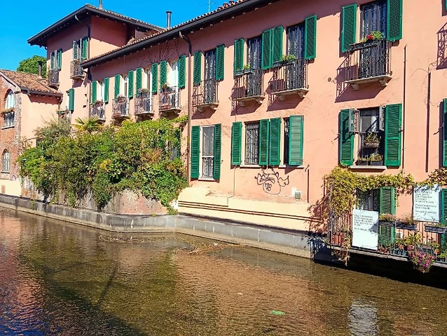 Pink canal-side architecture with green shutters and lush climbing vegetation in sunlight.