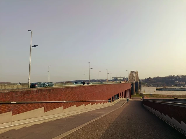 A modern brick ramp leads toward a bridge over the flowing river.