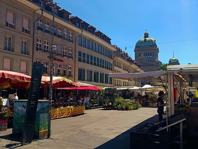 Market stalls with colorful awnings line a sunny urban plaza under clear skies.