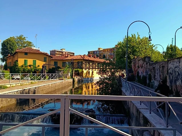 The observer views a sunlit canal and bridge from a metal railing.