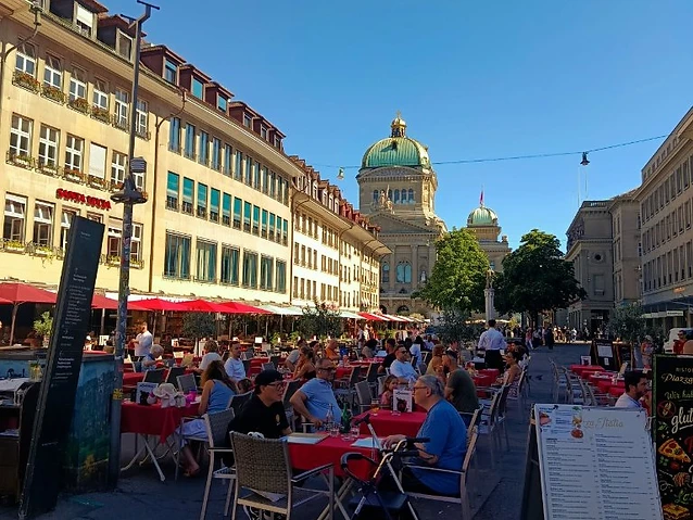 Dining area with red tables in a lively city square facing a dome.