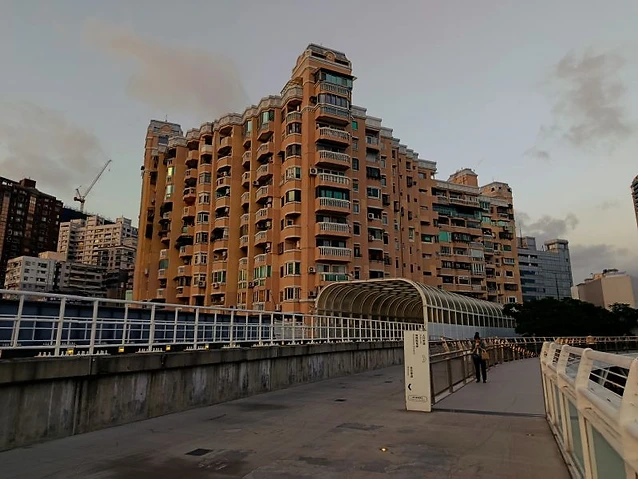 The observer walks along a modern pedestrian bridge with a large residential complex background.
