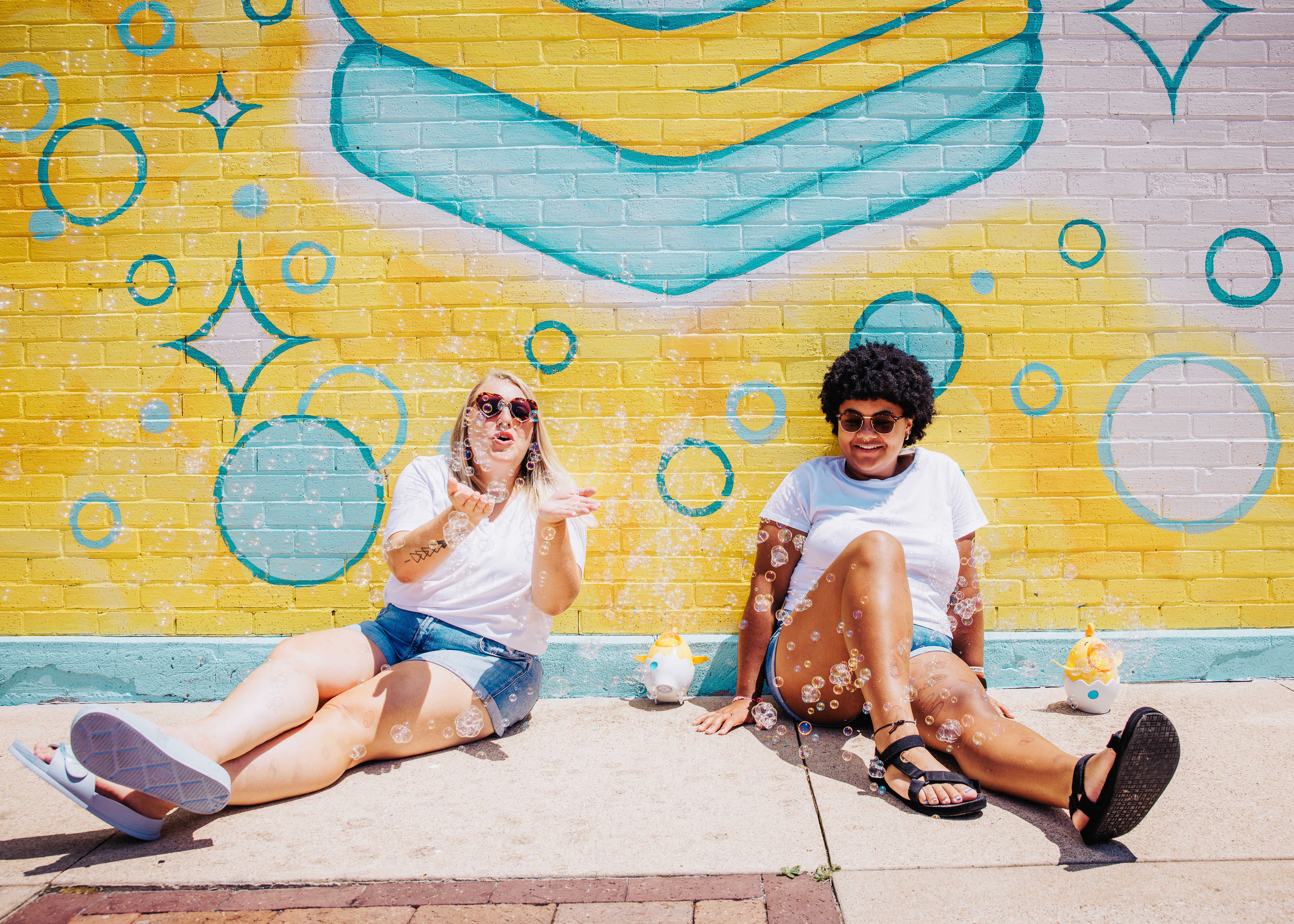 Cortney and Keeley in white shirts and sunglasses sitting in front of the colorful yellow Two Chicks Fluff & Fold mural