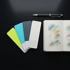 Illustration stylisée d’un nuancier de cartes vierges, d’un carnet ouvert aux taches de couleur et d’un stylo sur une table sombre
