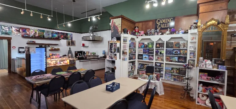 Wide view of Gaming Galley at dusk with warm amber interior glow, shelves of board games, wooden tables with dice and cards