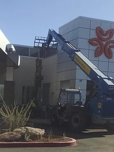 Blue telehandler extending its boom to install a braced saguaro at a modern commercial building entrance