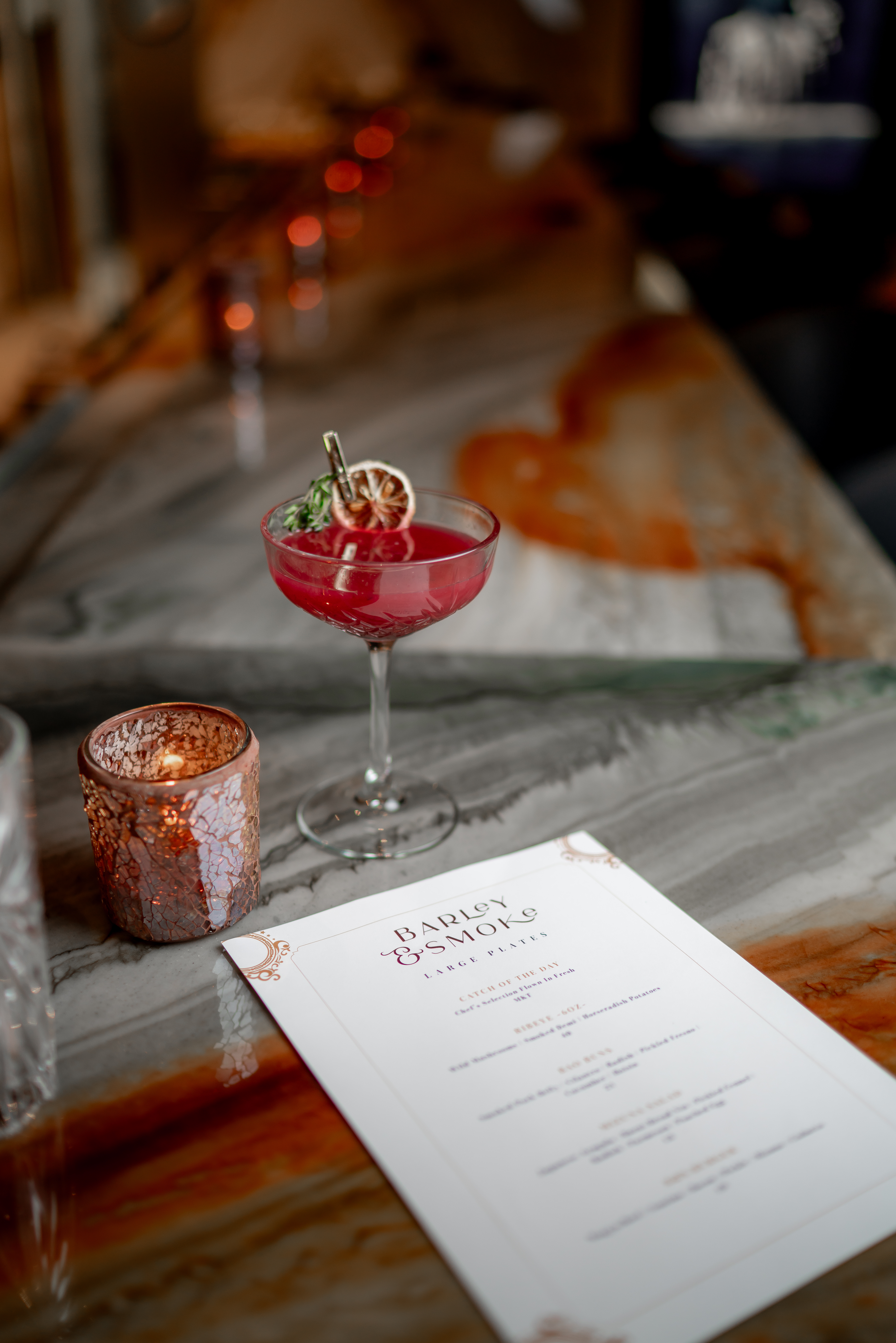 A craft cocktail and candle beside the Barley & Smoke menu