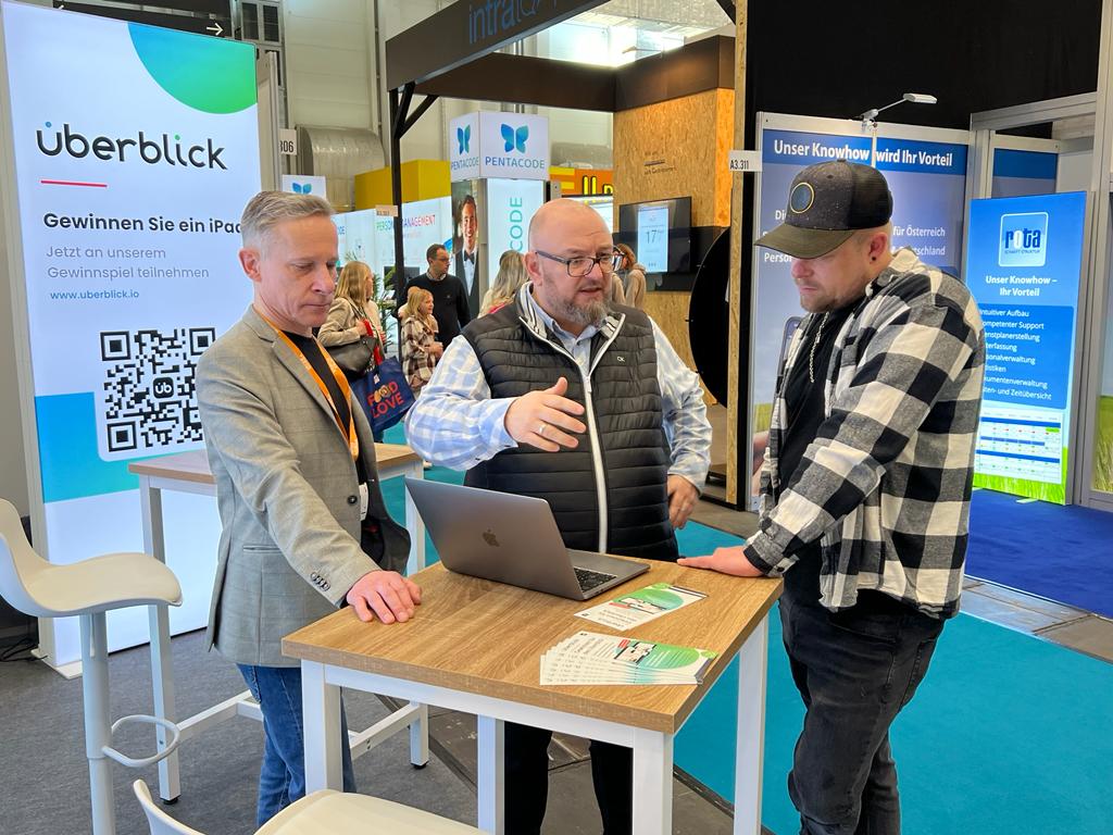 Drei Männer am Messestand um einen Stehtisch mit Laptop – Szene, die den Unterschied zwischen passiver Standwache und aktiver Kundengewinnung am Stand veranschaulicht