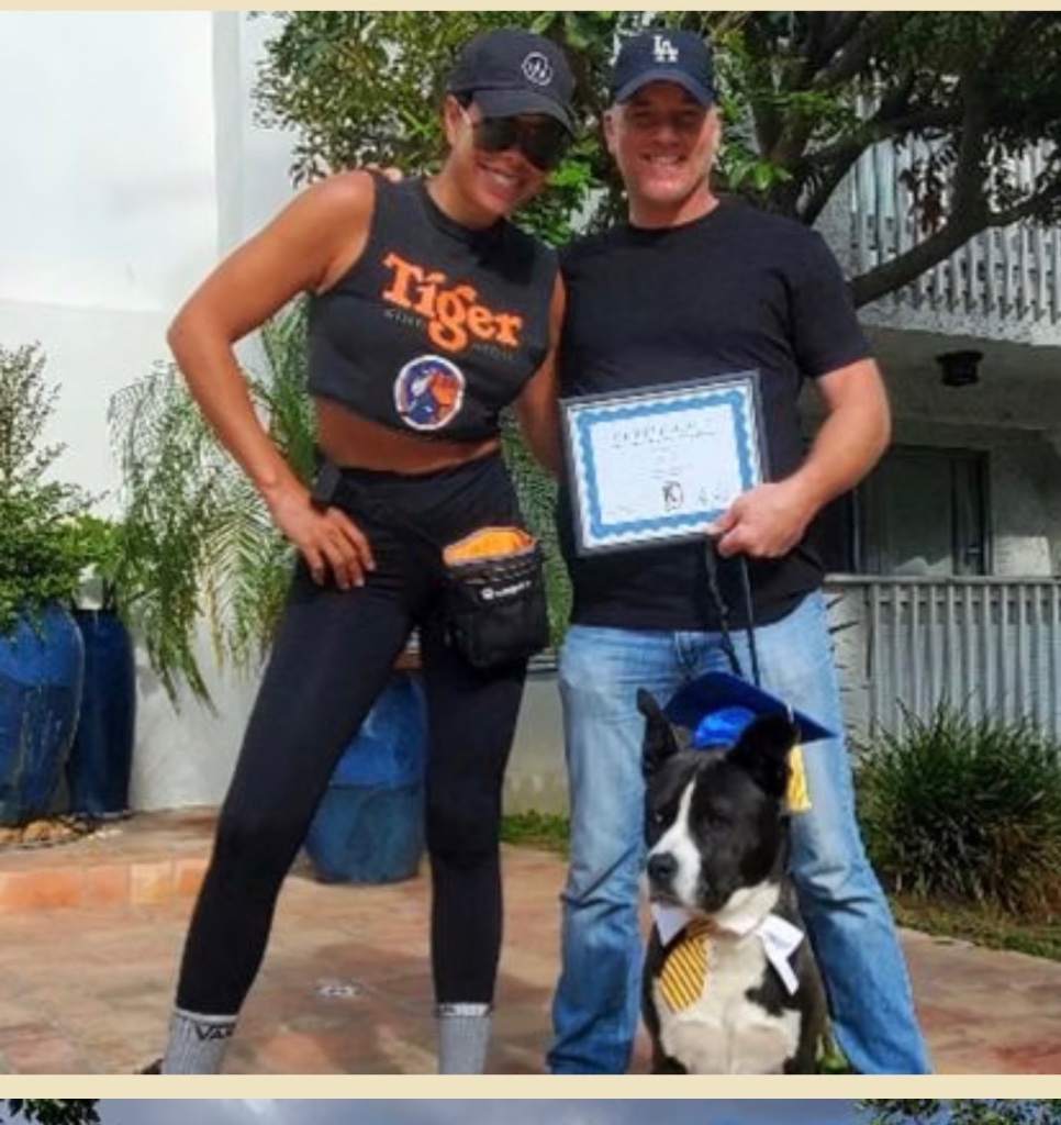A couple standing outside their home with their dog, who wears a graduation cap and tie, beside a Certificate of Completion.