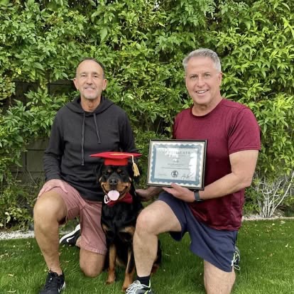 Two men kneeling with a Rottweiler in a red graduation cap.