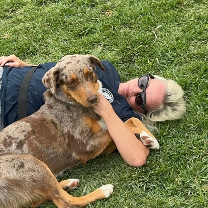 Johanna lies back in the grass with Ava, a merle-coated dog resting against her chest, both at ease in the afternoon light.