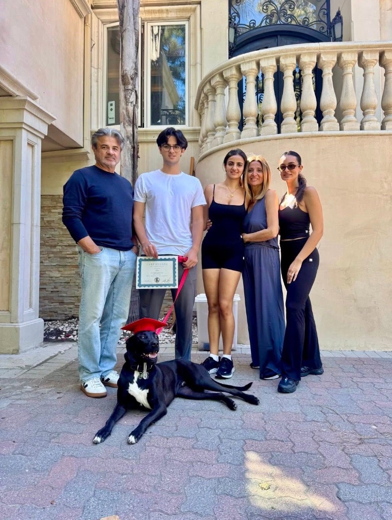 A family of five with their dog at graduation, the dog wearing a red cap and a certificate in hand.