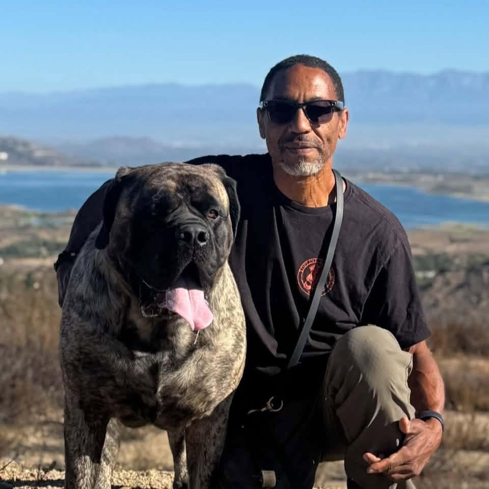 Illya kneels beside a large brindle dog against an open landscape.