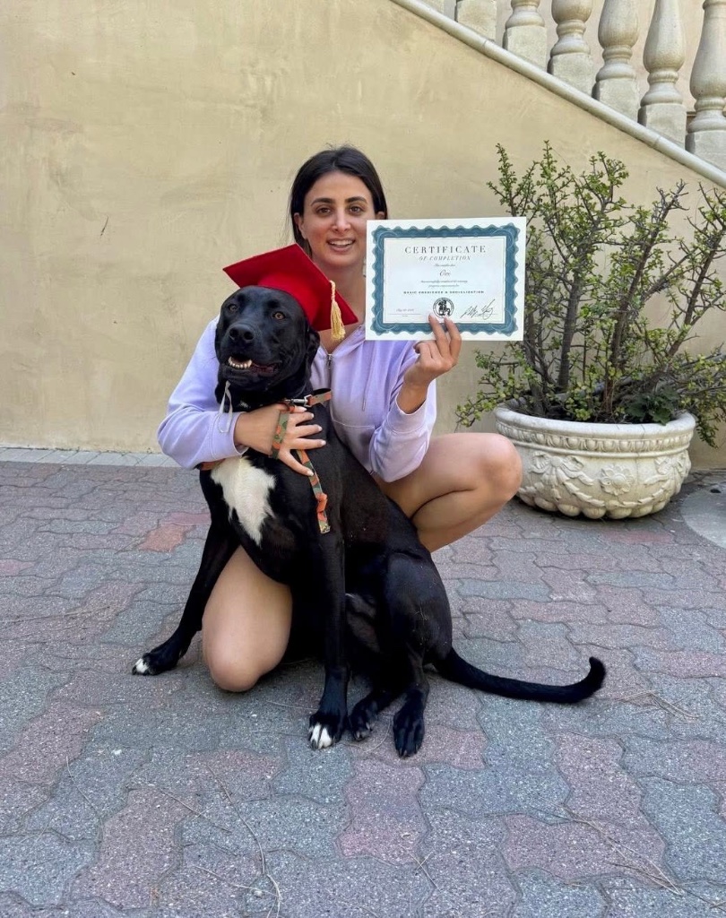 A woman kneeling with her black dog in a red graduation cap, holding a certificate.