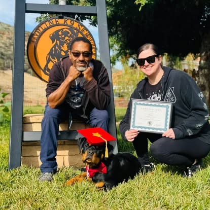 A couple kneeling with a Rottweiler puppy in a red graduation cap, in front of the Topline K9 sign.