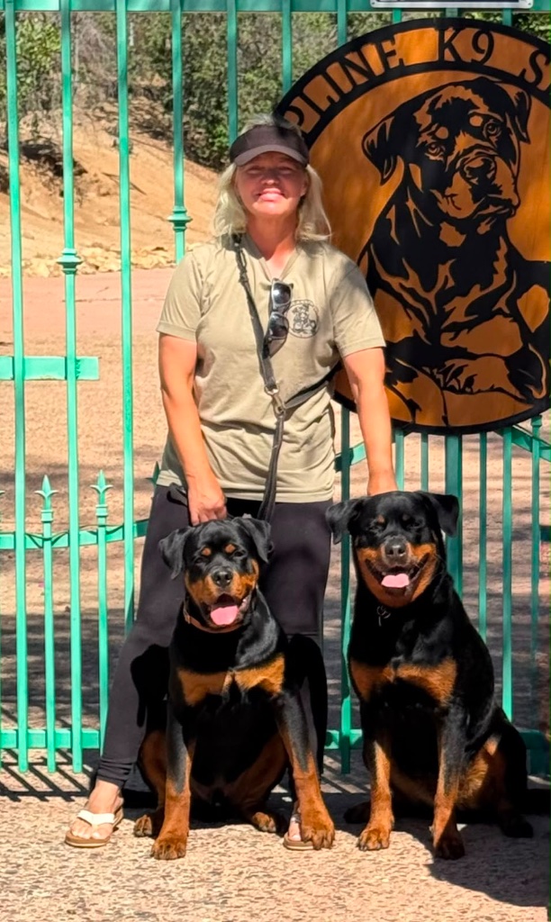 Johanna stands before a turquoise fence with two Rottweilers seated at her side.