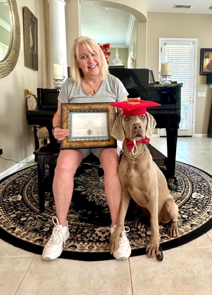 A woman seated beside her dog in a graduation cap, holding a framed certificate.