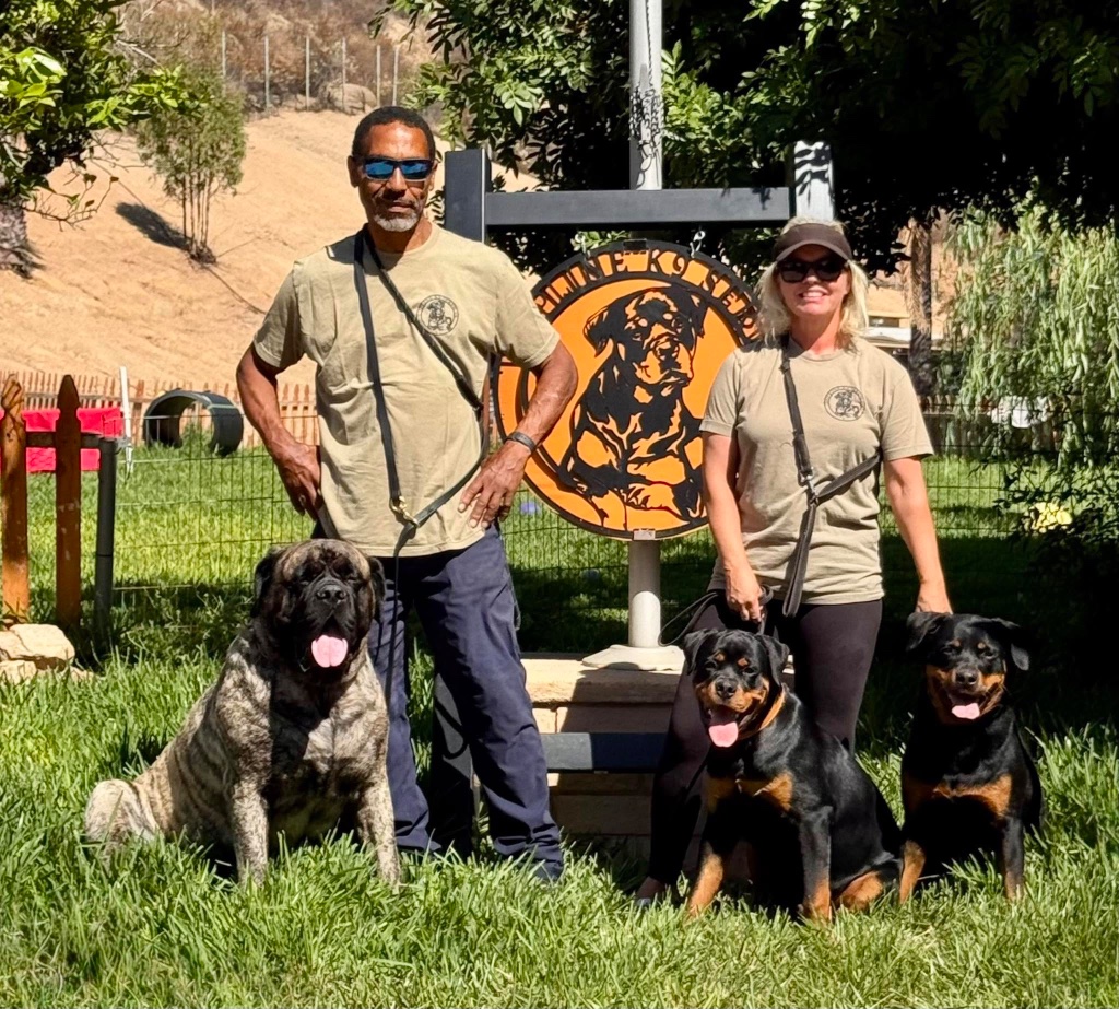 Illya and Johanna Hardy with three of their dogs, standing in front of the Topline K9 Services sign.