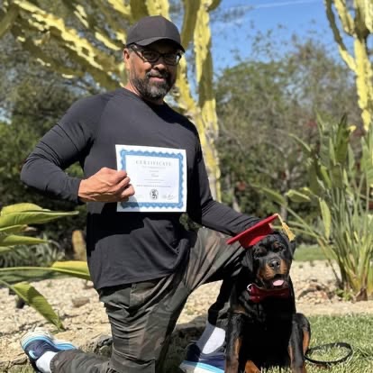 A man kneeling with a Rottweiler wearing a red graduation cap and bow tie.