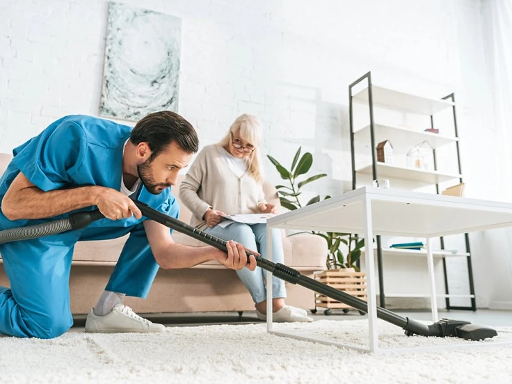 A caregiver in blue scrubs vacuuming under a coffee table while a senior woman sits on the couch nearby
