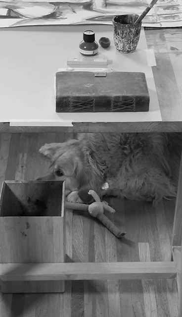 An artist's wooden desk with paper and brushes, and a golden retriever resting on the floor below