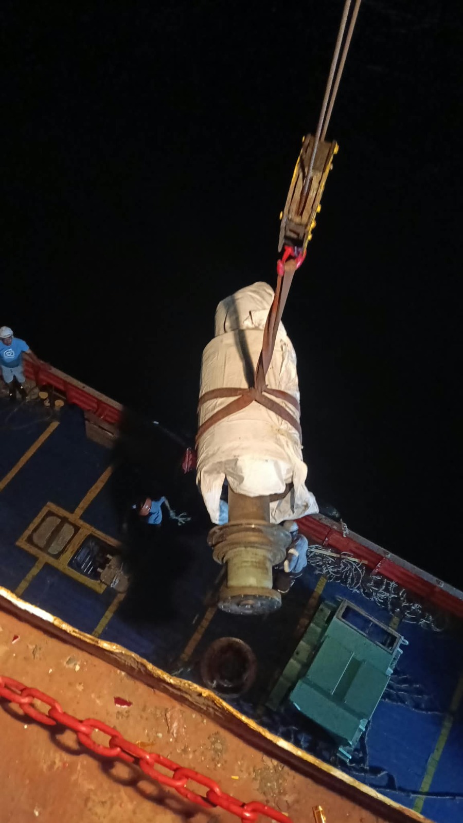 Crane lifting wrapped cylindrical machinery on the vessel deck with crew coordinating below