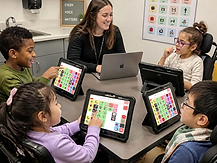 A speech-language pathologist sits at a table with four children using AAC tablets, several in wheelchairs, in a clinical setting.