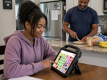 A teenager uses her AAC tablet to communicate about a snack with her father in the kitchen.
