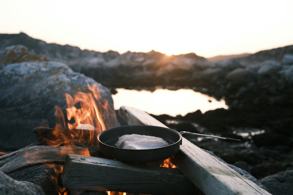 Raw fish fillet cooking in a black pan over a campfire at sunset on a rocky shoreline