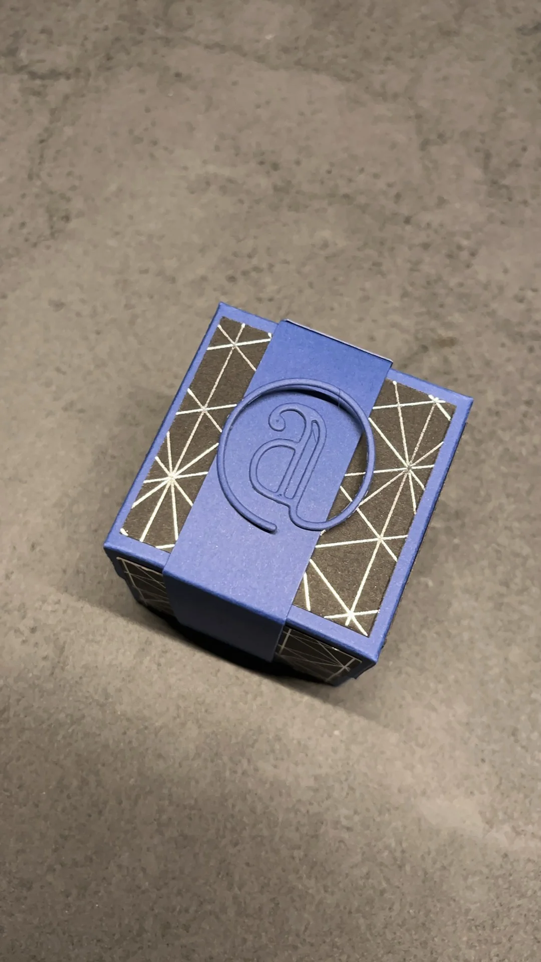 Boîte artisanale bleue au papier graphique noir et argenté, fermée par une bande monogrammée