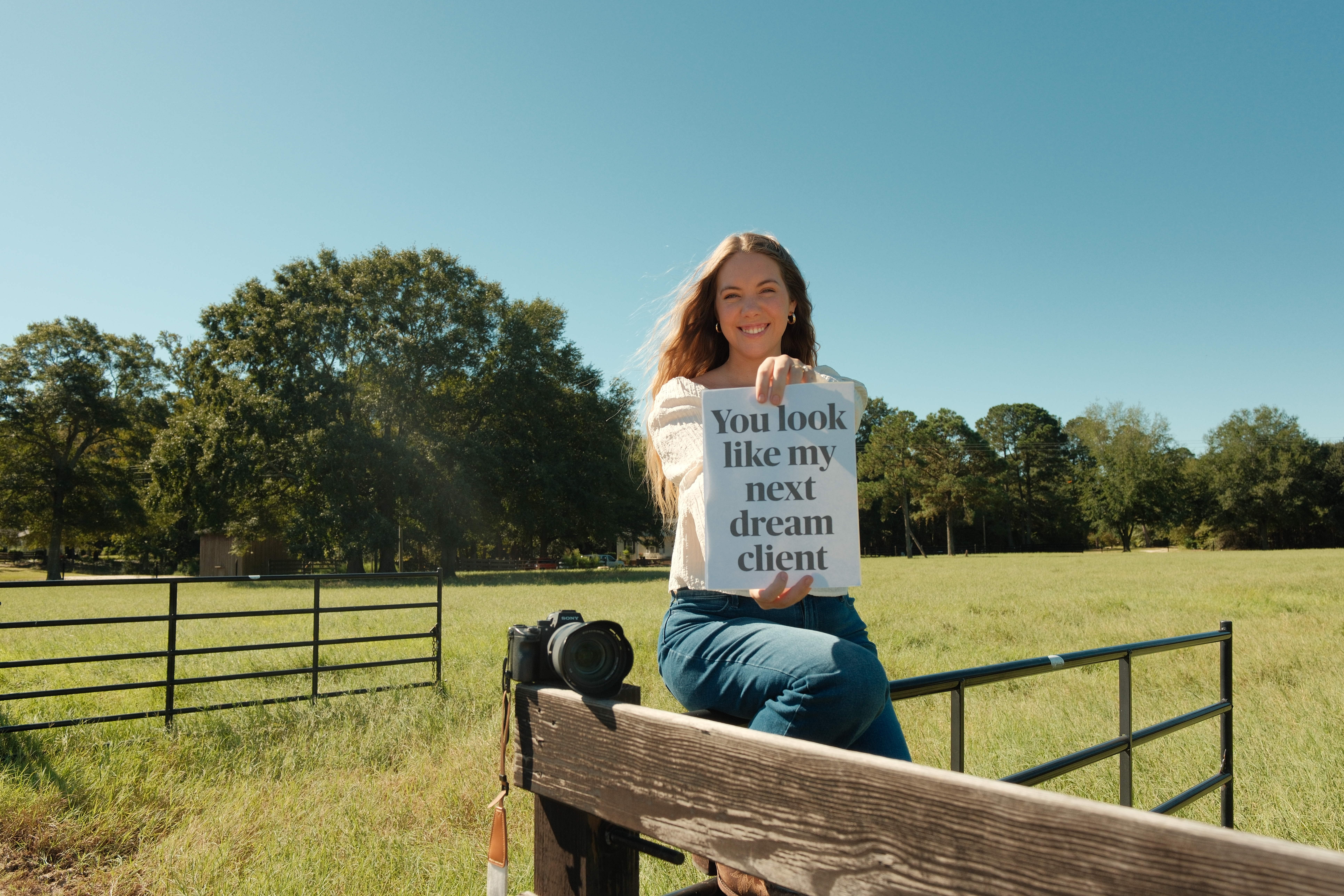 Emma sitting on a wooden fence in a meadow, holding a sign that reads You look like my next dream client