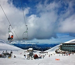 Ski lift carrying skiers at Parnassos Ski Centre with snow-covered mountains and chalet under a blue sky