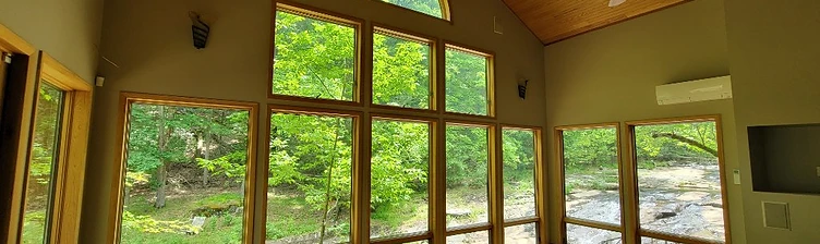 Sun-filled room with tall wood-framed windows looking onto the creek and forest