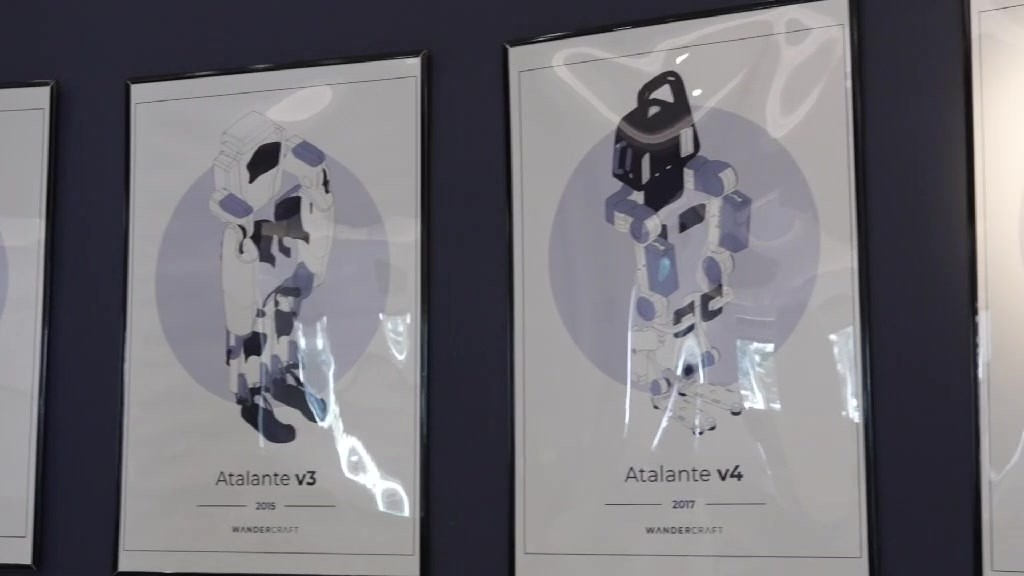Two framed posters on a dark wall showing illustrated Atalante v3 (2015) and Atalante v4 (2017) exoskeletons