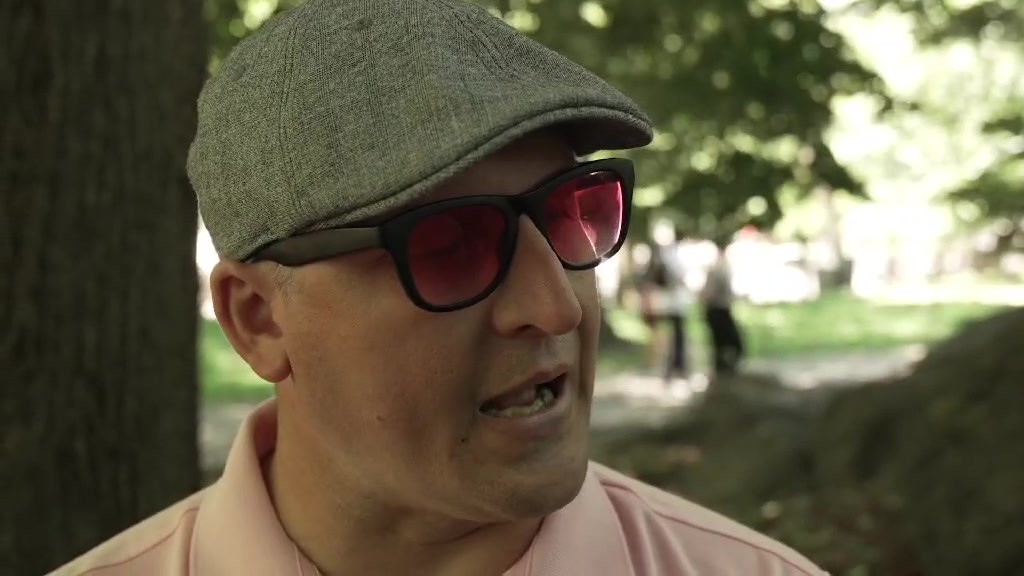 Joshua Holm, in a gray flat cap, tinted sunglasses and a pink polo, speaks outdoors under trees