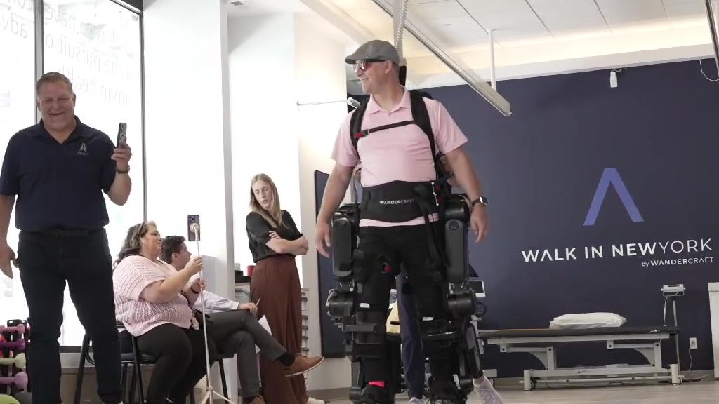 Joshua Holm walks in a Wandercraft exoskeleton at Walk In New York as staff watch