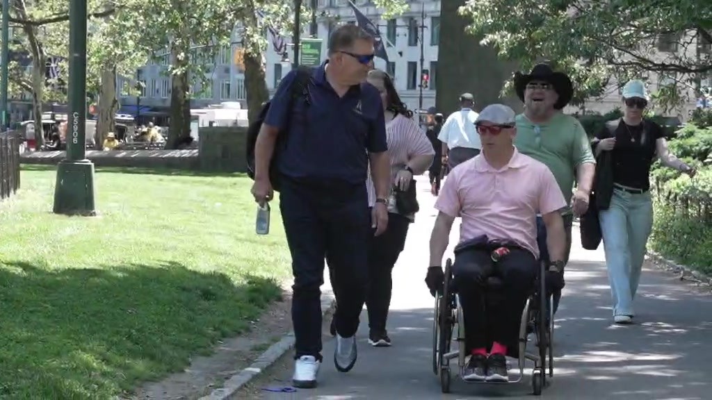 Joshua Holm in his wheelchair moves along a sunny city park path with Glenn Baker in a black cowboy hat and other supporters walking beside him
