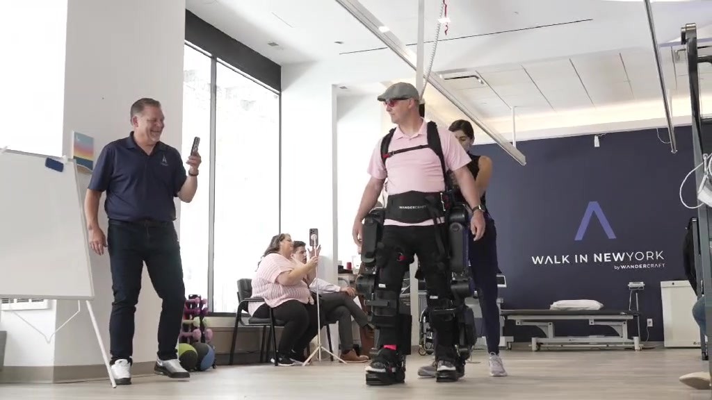 Joshua Holm walks upright in a Wandercraft exoskeleton across the Walk In New York clinic floor, a clinician behind him and onlookers filming