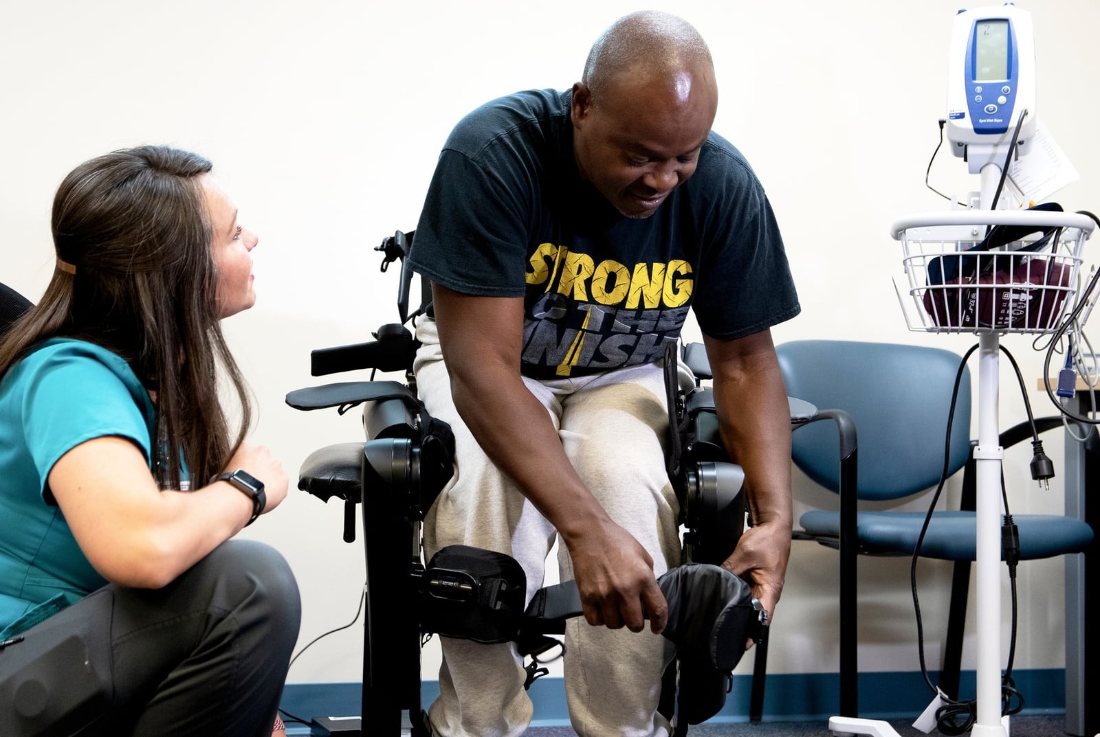 An occupational therapist kneels beside an Army veteran as he is fitted into an exoskeleton at a VA medical center