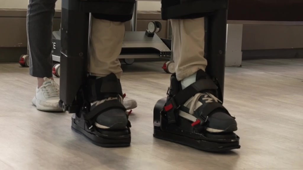 Close-up of a patient's feet strapped into the black footplates of an exoskeleton on a wood floor