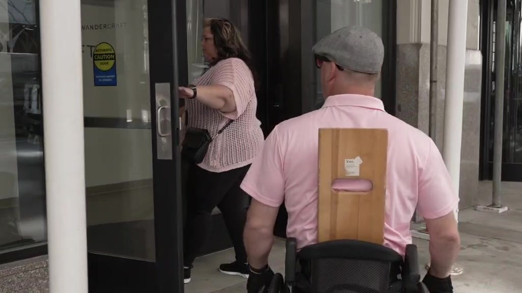 Joshua Holm, seen from behind in a pink polo and gray flat cap, arrives in his wheelchair at the glass doors of Walk In New York as a woman holds the door