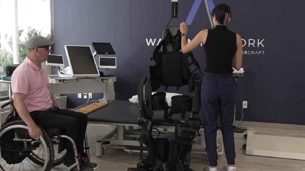 Joshua Holm sits in his wheelchair beside a Wandercraft exoskeleton hanging from an overhead support as a clinician adjusts it